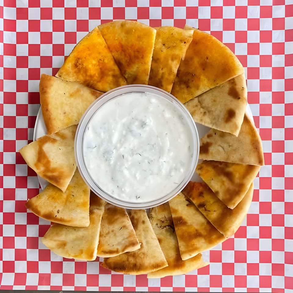 Pita bread triangles arranged around a container of tzatziki sauce on a red and white checkered background.