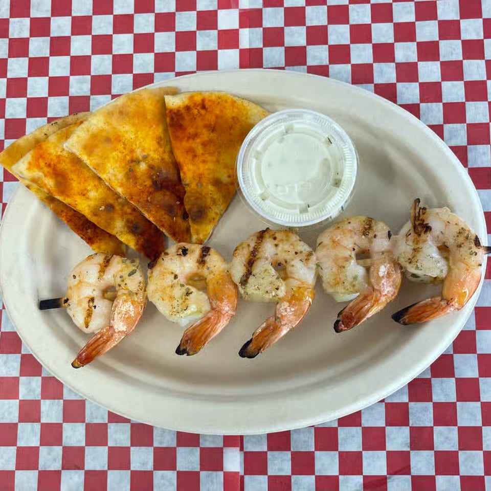 Grilled shrimp skewers, pita bread, and a dipping sauce on a white plate over a red and white checkered tablecloth.