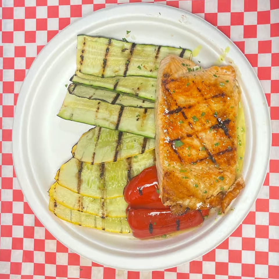 Grilled salmon with zucchini and red peppers on a white plate, on a red and white checkered tablecloth.